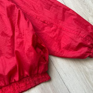May include: A close-up of a bright red jacket. The jacket has a shiny, crinkled texture. The sleeves and bottom hem are elasticized. The jacket appears to be made of a lightweight material.