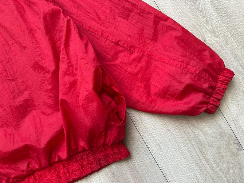 May include: A close-up of a bright red jacket. The jacket has a shiny, crinkled texture. The sleeves and bottom hem are elasticized. The jacket appears to be made of a lightweight material.