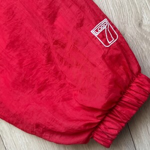 May include: Close-up of a red nylon jacket sleeve. The sleeve has a white logo patch and a ribbed cuff. The fabric has a slight sheen and a crinkled texture.