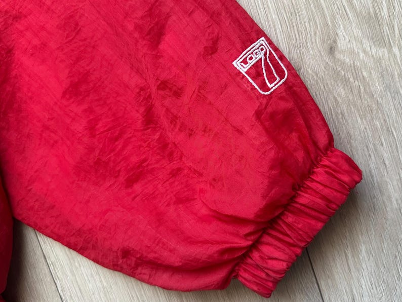 May include: Close-up of a red nylon jacket sleeve. The sleeve has a white logo patch and a ribbed cuff. The fabric has a slight sheen and a crinkled texture.