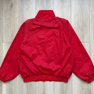 May include: A bright red nylon jacket with a high collar and elastic cuffs and waistband. The jacket is a solid color and appears to be lightweight. The jacket is laid flat on a light wood surface.