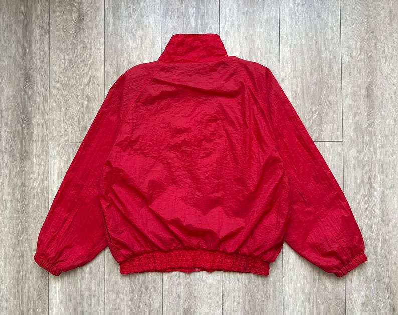 May include: A bright red nylon jacket with a high collar and elastic cuffs and waistband. The jacket is a solid color and appears to be lightweight. The jacket is laid flat on a light wood surface.