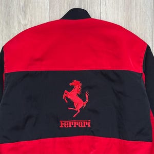May include: Red and black jacket featuring the Ferrari logo. The jacket has a red upper section and black lower section with the Ferrari logo embroidered in red. The jacket is made of a smooth, possibly nylon, material.