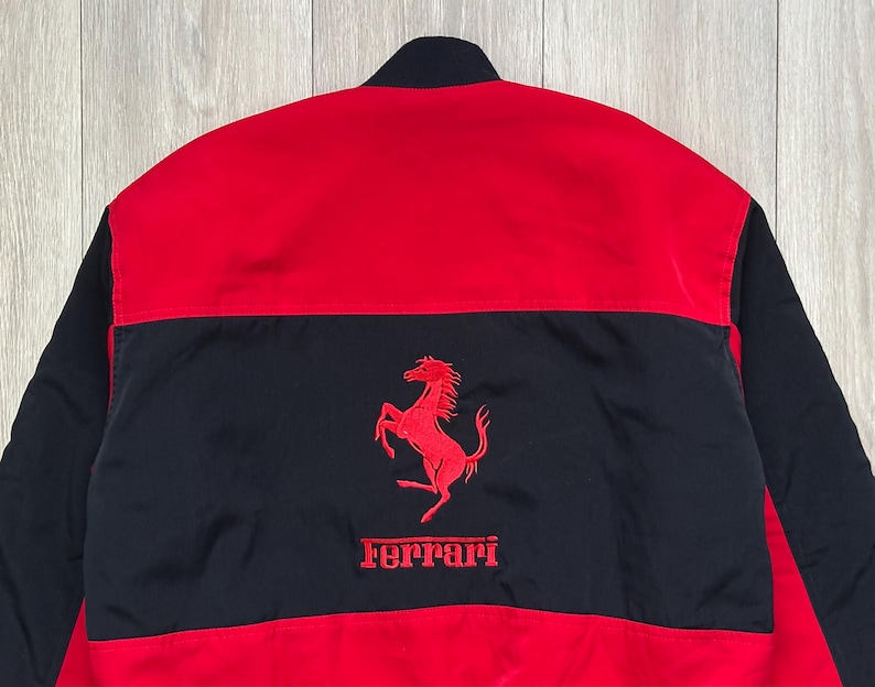 May include: Red and black jacket featuring the Ferrari logo. The jacket has a red upper section and black lower section with the Ferrari logo embroidered in red. The jacket is made of a smooth, possibly nylon, material.