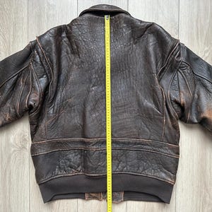 May include: A dark brown leather bomber jacket with a distressed texture, long sleeves, and a ribbed hem. A yellow measuring tape is placed vertically down the centre back, showing a length of approximately 66 cm from the collar to the hem.