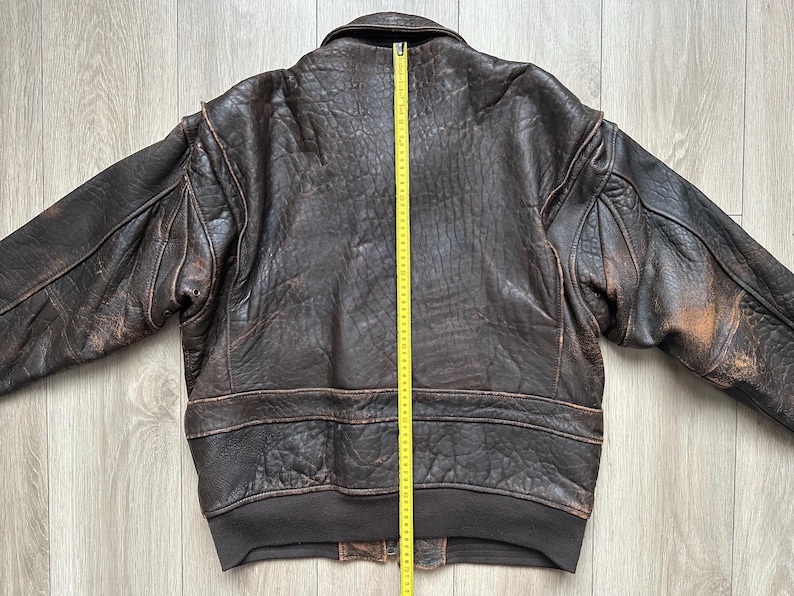 May include: A dark brown leather bomber jacket with a distressed texture, long sleeves, and a ribbed hem. A yellow measuring tape is placed vertically down the centre back, showing a length of approximately 66 cm from the collar to the hem.