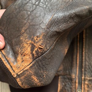 May include: A dark brown leather jacket featuring a heavily distressed and worn texture with visible surface abrasion and scuffing on the sleeve. The jacket includes a dark brown ribbed knit cuff and a central zipper closure with matching leather panels.