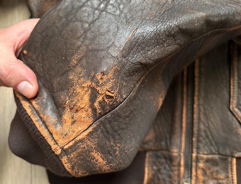 May include: A dark brown leather jacket featuring a heavily distressed and worn texture with visible surface abrasion and scuffing on the sleeve. The jacket includes a dark brown ribbed knit cuff and a central zipper closure with matching leather panels.