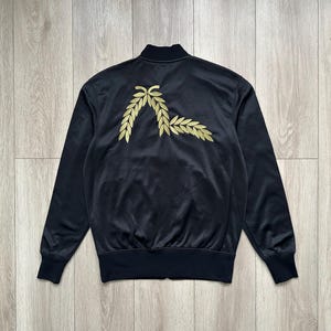 May include: Black track jacket with a gold laurel wreath design on the back. The jacket has a stand-up collar, long sleeves, and ribbed cuffs and hem. The jacket is made of a smooth, shiny material.