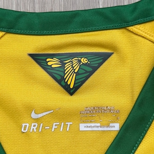 Pu&ograve; includere: Primo piano di una maglia sportiva gialla Dri-FIT con finiture verdi. Un emblema triangolare con un disegno di uccello giallo &egrave; centrato sul petto. Il logo Nike e "DRI-FIT" sono visibili. La maglia &egrave; prodotta in Thailandia.