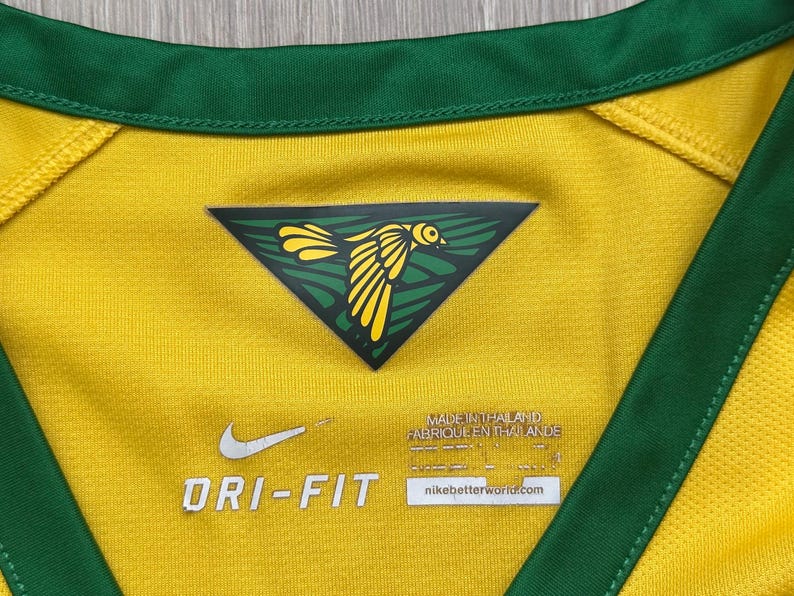 Pu&ograve; includere: Primo piano di una maglia sportiva gialla Dri-FIT con finiture verdi. Un emblema triangolare con un disegno di uccello giallo &egrave; centrato sul petto. Il logo Nike e "DRI-FIT" sono visibili. La maglia &egrave; prodotta in Thailandia.