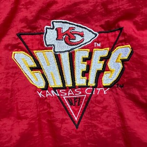May include: Red jacket with the Kansas City Chiefs logo embroidered on the front. The logo features the team name in yellow and white, with a red arrowhead and the letters "KC". The words "Kansas City" are also embroidered.