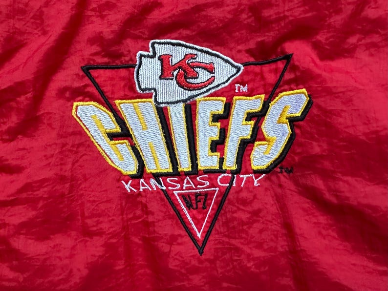 May include: Red jacket with the Kansas City Chiefs logo embroidered on the front. The logo features the team name in yellow and white, with a red arrowhead and the letters "KC". The words "Kansas City" are also embroidered.