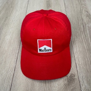 Peut inclure: Une casquette de baseball rouge vif dotée d'un écusson rectangulaire sur le panneau avant. L'écusson affiche le logo Marlboro blanc et rouge avec le nom de la marque écrit en noir. La casquette présente une visière incurvée et une calotte structurée.
