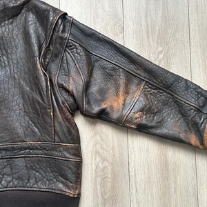 May include: A dark brown leather bomber jacket featuring a heavily textured, distressed finish with visible lighter tan wear patterns on the sleeve. The garment includes a dark brown ribbed knit cuff and a matching ribbed hem at the waistline.