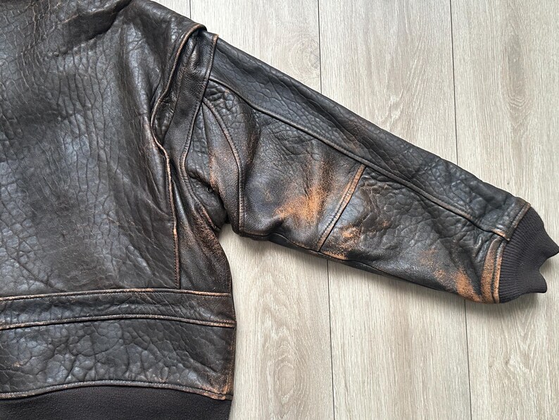 May include: A dark brown leather bomber jacket featuring a heavily textured, distressed finish with visible lighter tan wear patterns on the sleeve. The garment includes a dark brown ribbed knit cuff and a matching ribbed hem at the waistline.