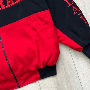 May include: A black and red jacket with the word "RACING" embroidered in red on the chest. The jacket has a full zipper and a pocket. The sleeves are black with red embroidered text. The cuffs and hem are black.