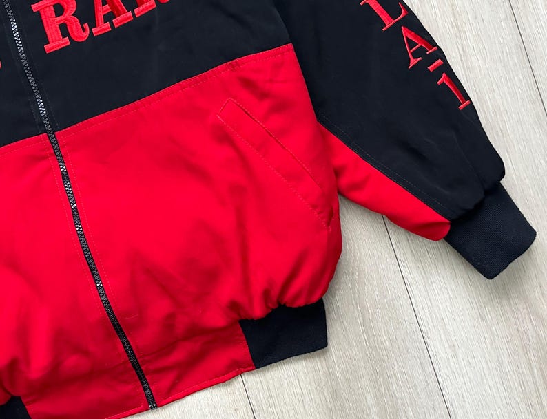 May include: A black and red jacket with the word "RACING" embroidered in red on the chest. The jacket has a full zipper and a pocket. The sleeves are black with red embroidered text. The cuffs and hem are black.