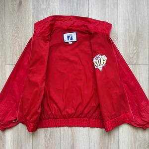May include: Bright red vintage jacket with a full zipper closure. The jacket has a white and gold logo on the left sleeve and a small patch on the chest. The jacket is open, revealing the interior lining.