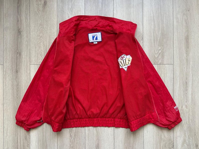 May include: Bright red vintage jacket with a full zipper closure. The jacket has a white and gold logo on the left sleeve and a small patch on the chest. The jacket is open, revealing the interior lining.