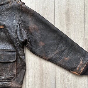 May include: A dark brown distressed leather bomber jacket featuring a textured finish, a fold-down collar, and a rectangular leather patch on the chest embossed with the text AVIREX LTD. The jacket includes a front flap pocket and ribbed cuffs.