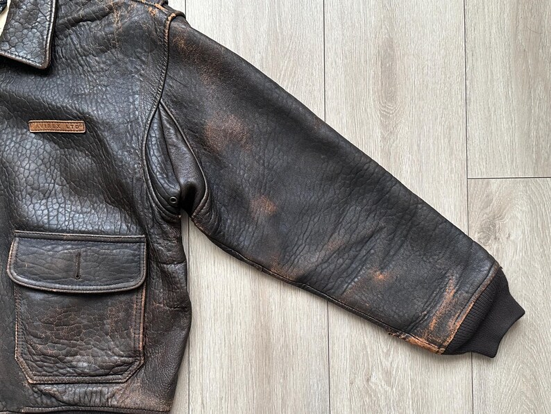 May include: A dark brown distressed leather bomber jacket featuring a textured finish, a fold-down collar, and a rectangular leather patch on the chest embossed with the text AVIREX LTD. The jacket includes a front flap pocket and ribbed cuffs.