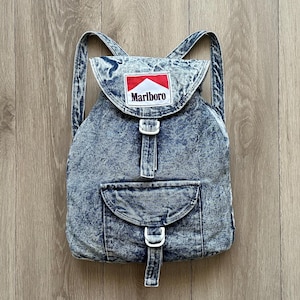 May include: A blue acid-wash denim backpack featuring a flap closure with a white, red, and black Marlboro logo patch. The bag includes two adjustable shoulder straps, a front pocket with a flap, and white plastic buckle hardware closures.