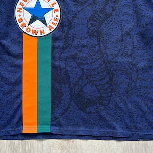 May include: A navy blue jersey with a patterned design and a vertical orange and green stripe. The jersey features a circular emblem with a blue star and the words "BROWN ALE". The collar and sleeve cuffs are green.