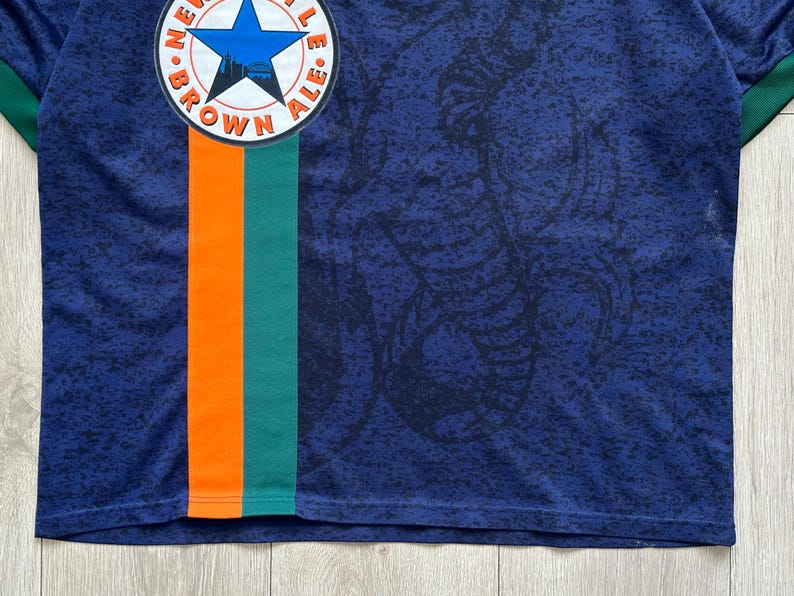 May include: A navy blue jersey with a patterned design and a vertical orange and green stripe. The jersey features a circular emblem with a blue star and the words "BROWN ALE". The collar and sleeve cuffs are green.