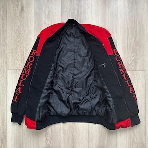 May include: Black and red Formula 1 jacket. The jacket has a black body with red accents on the shoulders and sleeves. The word "FORMULA 1" is printed vertically in red on both sleeves. The jacket is open, revealing a black quilted lining.