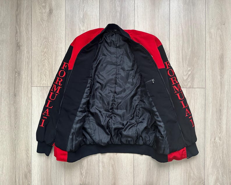 May include: Black and red Formula 1 jacket. The jacket has a black body with red accents on the shoulders and sleeves. The word "FORMULA 1" is printed vertically in red on both sleeves. The jacket is open, revealing a black quilted lining.