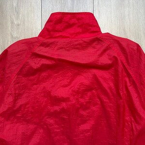 May include: A close-up of a bright red jacket, showcasing the high collar and the texture of the fabric. The jacket appears to be made of a lightweight, possibly nylon or polyester material, with a slight sheen. The focus is on the upper portion of the garment.