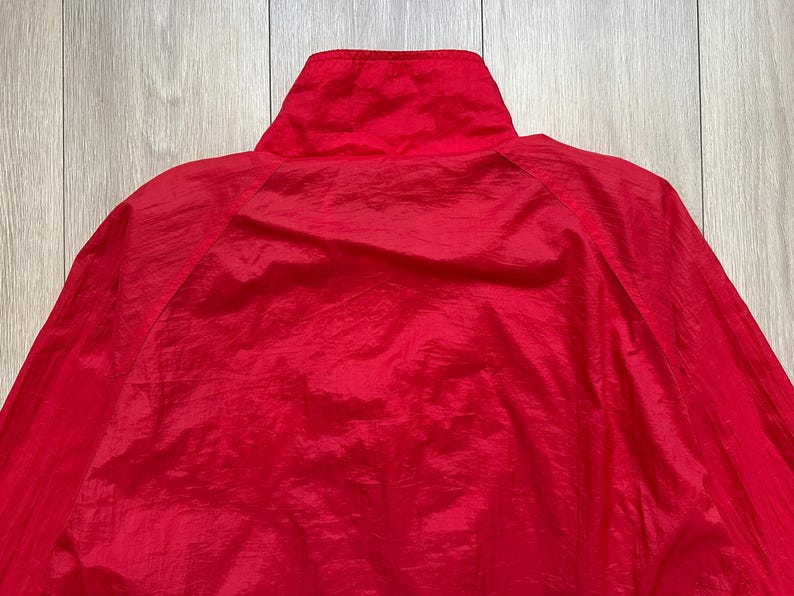 May include: A close-up of a bright red jacket, showcasing the high collar and the texture of the fabric. The jacket appears to be made of a lightweight, possibly nylon or polyester material, with a slight sheen. The focus is on the upper portion of the garment.