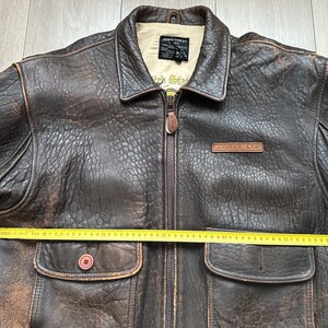 May include: A dark brown distressed leather jacket with a front zip, two flap pockets, and a beige interior lining. A yellow measuring tape is placed across the chest, showing a width of approximately 61 cm. The label reads JACKET TYPE G1.