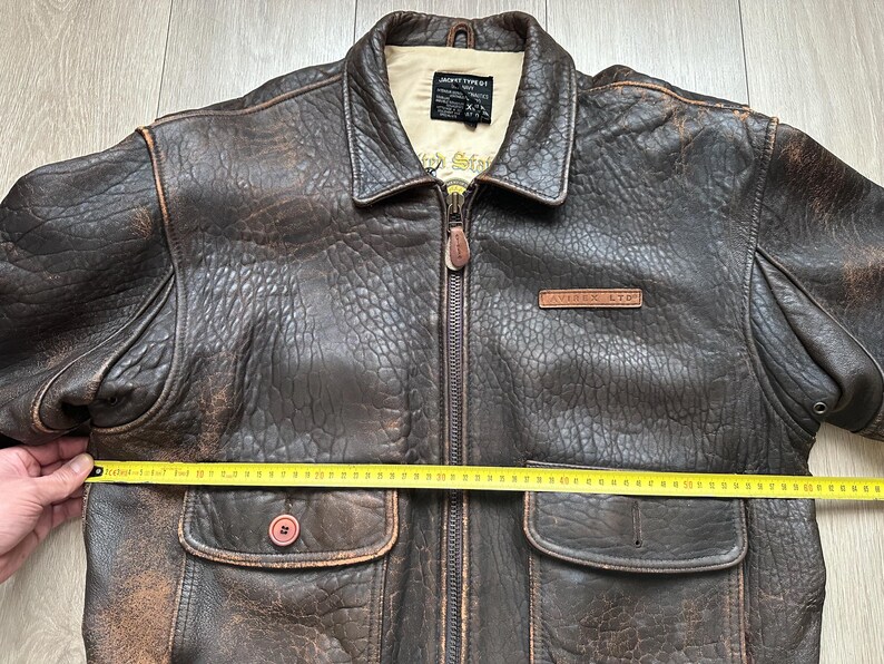 May include: A dark brown distressed leather jacket with a front zip, two flap pockets, and a beige interior lining. A yellow measuring tape is placed across the chest, showing a width of approximately 61 cm. The label reads JACKET TYPE G1.