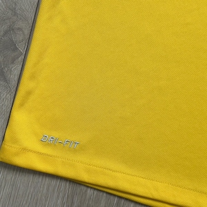 Pu&ograve; includere: Primo piano di una maglietta sportiva giallo brillante. La maglietta ha un tessuto strutturato e le parole "DRI-FIT" sono ricamate in argento sul lato inferiore sinistro. La maglietta &egrave; realizzata in un materiale leggero e traspirante.