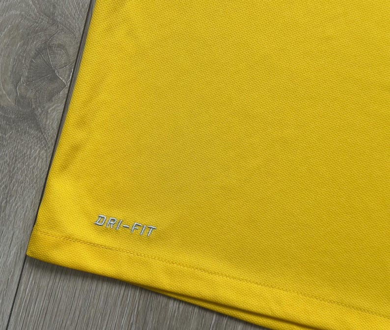 Pu&ograve; includere: Primo piano di una maglietta sportiva giallo brillante. La maglietta ha un tessuto strutturato e le parole "DRI-FIT" sono ricamate in argento sul lato inferiore sinistro. La maglietta &egrave; realizzata in un materiale leggero e traspirante.