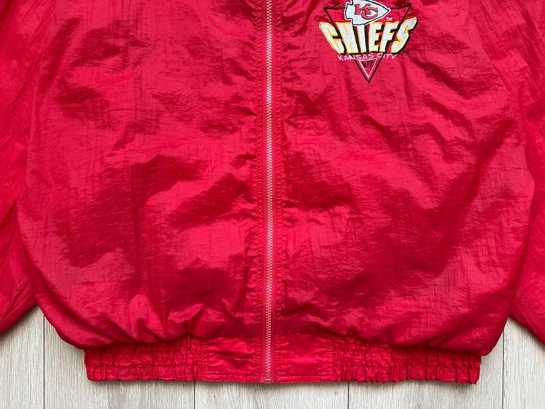 May include: A red nylon jacket with a full zipper and elastic cuffs and waist. The jacket features the Kansas City Chiefs logo on the upper left chest.