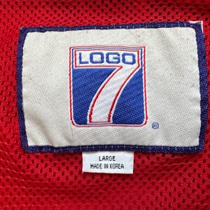 May include: Close-up of a red mesh fabric with a white label featuring the text "LOGO 7" in a blue and red design. The label also includes the text "LARGE" and "MADE IN KOREA".