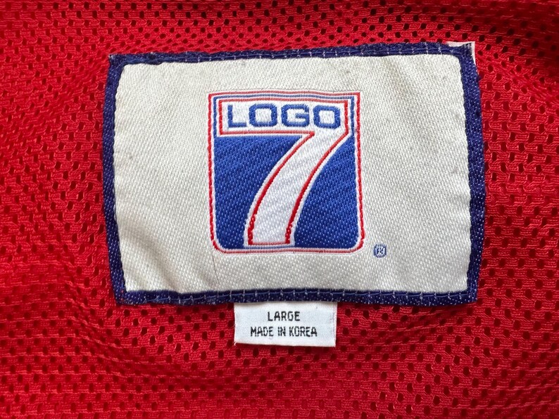 May include: Close-up of a red mesh fabric with a white label featuring the text "LOGO 7" in a blue and red design. The label also includes the text "LARGE" and "MADE IN KOREA".