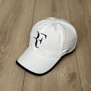 Può includere: Cappellino da baseball bianco con bordo nero e logo RF ricamato sul davanti. Lo swoosh Nike è visibile sul lato. Il cappellino è realizzato in materiale leggero.
