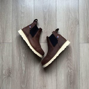 May include: A pair of brown leather Chelsea boots with white soles. The boots have a pull-on design and a small American flag logo on the side.