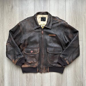 May include: A dark brown distressed leather bomber jacket featuring a front zip fastening, a fold-down collar, and two large flap pockets. The jacket has dark brown ribbed cuffs and a hem, with a cream-coloured interior lining and a visible label.