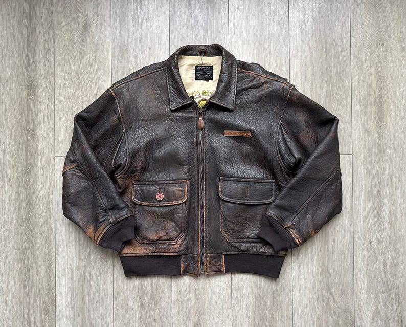 May include: A dark brown distressed leather bomber jacket featuring a front zip fastening, a fold-down collar, and two large flap pockets. The jacket has dark brown ribbed cuffs and a hem, with a cream-coloured interior lining and a visible label.