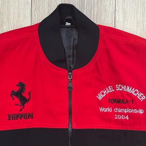 May include: Red and black zip-up jacket featuring the Ferrari logo and the words "MICHAEL SCHUMACHER FORMULA-1 World championship 2004" embroidered in white. The jacket has a black collar and a full-length zipper.