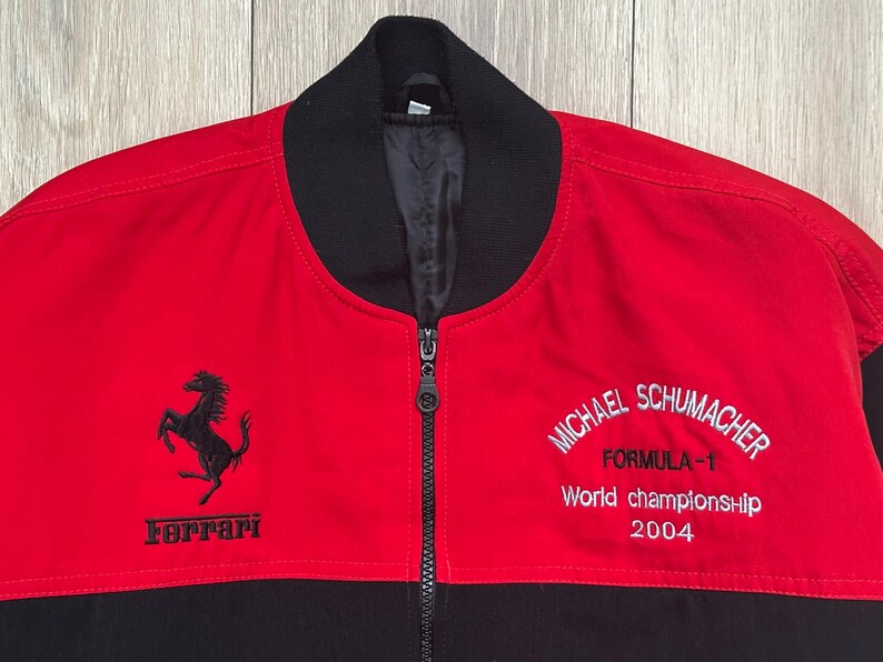 May include: Red and black zip-up jacket featuring the Ferrari logo and the words "MICHAEL SCHUMACHER FORMULA-1 World championship 2004" embroidered in white. The jacket has a black collar and a full-length zipper.