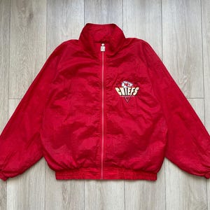 May include: A red zip-up jacket with the Kansas City Chiefs logo on the chest. The jacket has a high collar and elastic cuffs and hem. The logo is in white, yellow, and red.