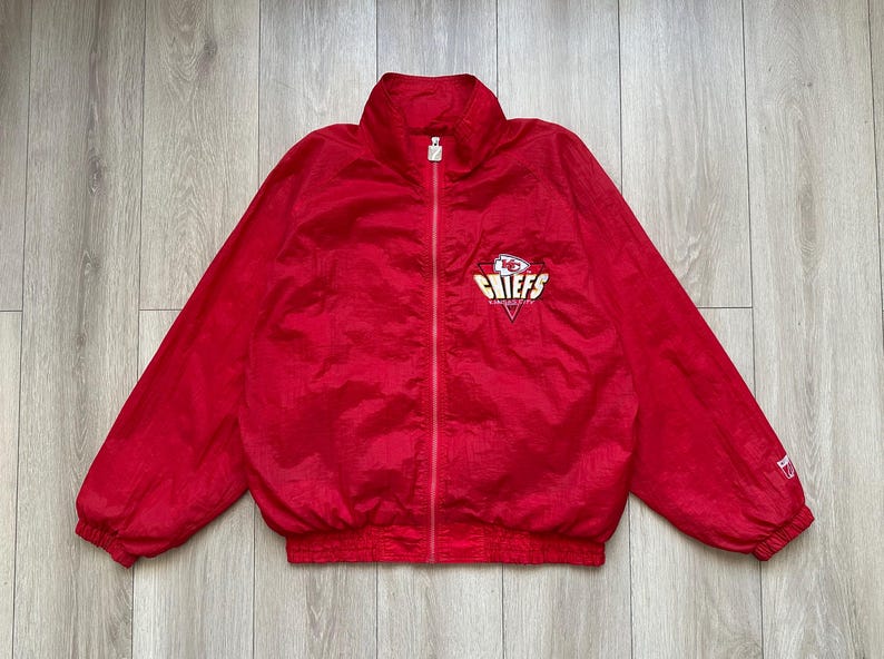 May include: A red zip-up jacket with the Kansas City Chiefs logo on the chest. The jacket has a high collar and elastic cuffs and hem. The logo is in white, yellow, and red.