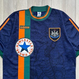 May include: Navy blue Adidas soccer jersey with green and orange accents. Features the Newcastle Brown Ale logo, the Adidas logo, and a team crest. The jersey has a textured pattern.