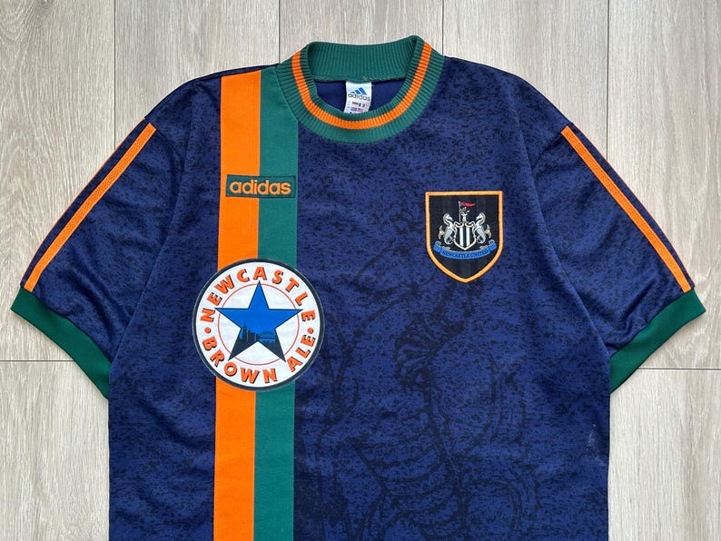 May include: Navy blue Adidas soccer jersey with green and orange accents. Features the Newcastle Brown Ale logo, the Adidas logo, and a team crest. The jersey has a textured pattern.